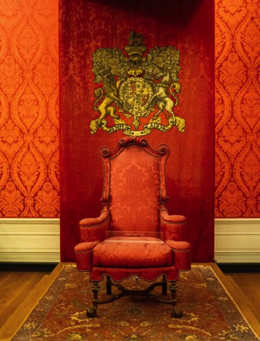 A chair of state at Kensington Palace under its crimson damask canopy, the gilded royal arms with lion and unicorn mounted on the wall above
