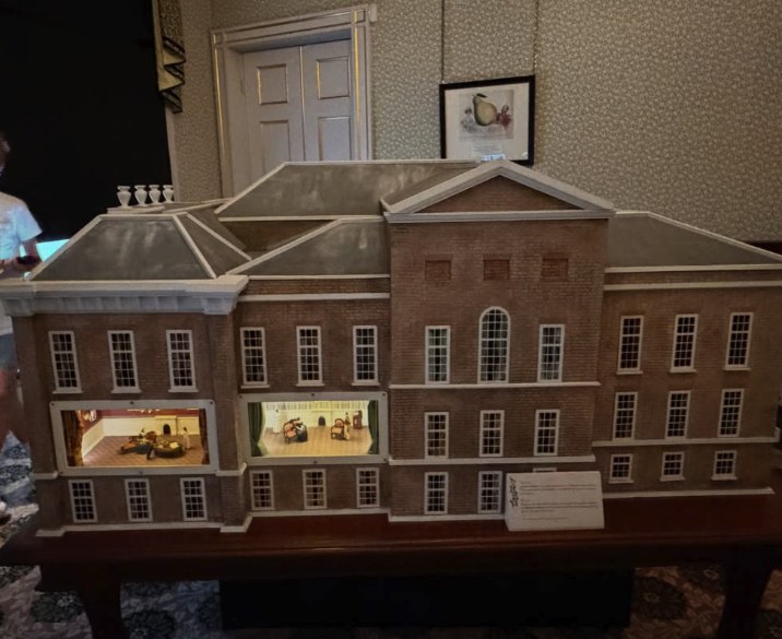 A large cutaway model of Kensington Palace on a table, two furnished rooms lit inside the brick south front