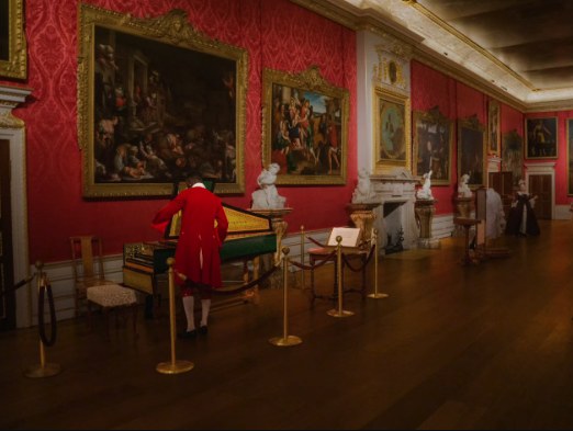 A green harpsichord in the King's Gallery with a costumed interpreter in an eighteenth-century red coat beside it, the long room stretching away past marble busts