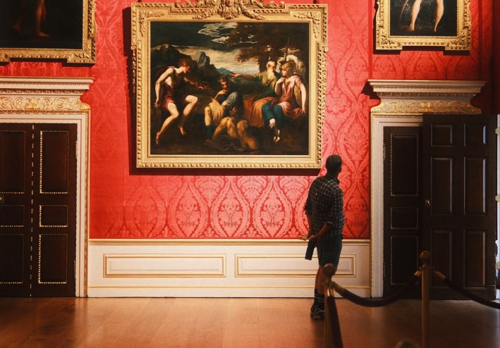 A lone visitor looking up at a large Old Master painting in the King's Gallery, the crimson damask running the length of the wall between panelled doors