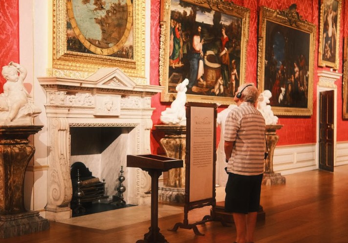 A visitor wearing the palace's audio-guide headset in the King's Gallery, standing before Old Master paintings hung on crimson damask beside a marble chimneypiece