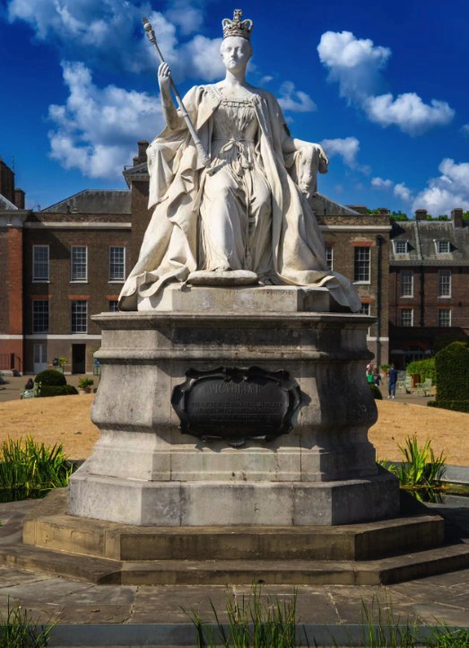 The Queen Victoria statue from the front, sceptre in hand on its carved stone plinth, with the palace and clipped hedging behind it