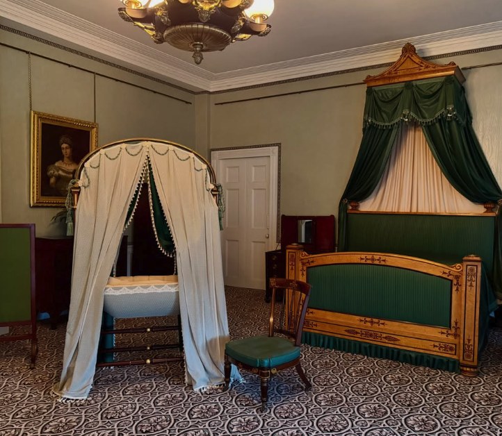 The bedroom in which Queen Victoria was born, presented with a satinwood bed under a green half-tester and a curtained cot standing beside it